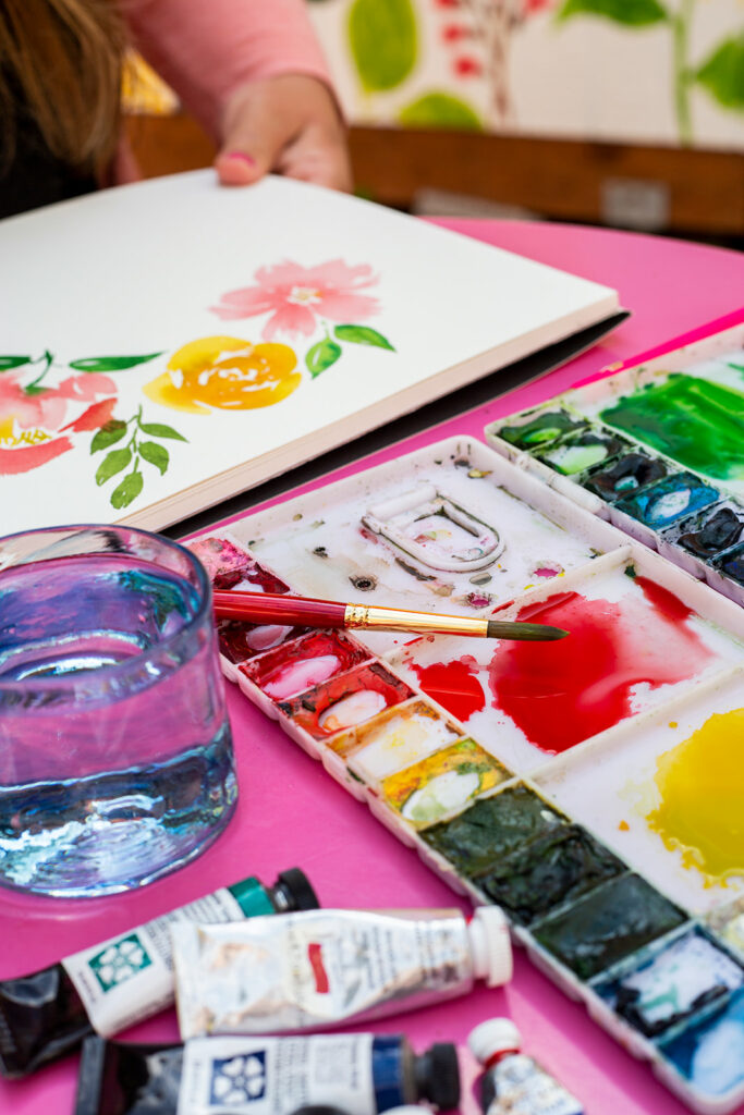 Watercolor paints and palette in the foreground with watercolor flowers in the background